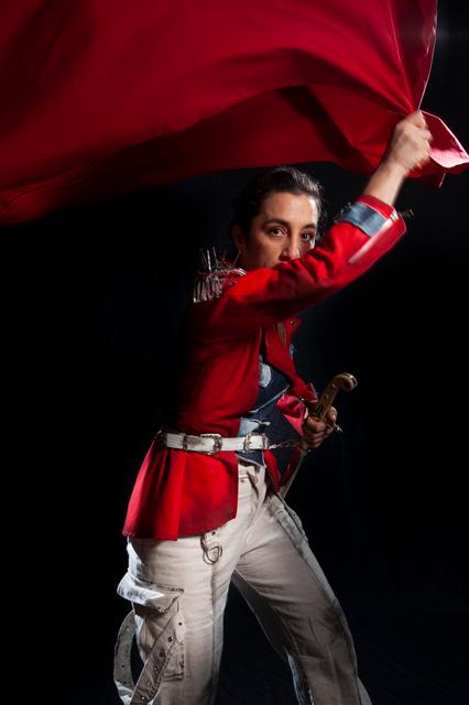 Una persona vestida con un traje rojo agita una bandera mientras sostiene una espada.