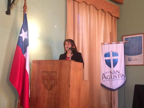 Una mujer habla desde un podio en un evento formal con banderas y un banner visible detrás.