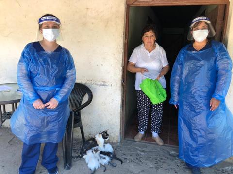 Tres personas, dos con trajes de protección y una mujer en la puerta de una casa, junto a un gato.