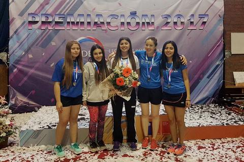 Un grupo de cinco chicas posando en un evento de premiación con medallas y un ramo de flores.