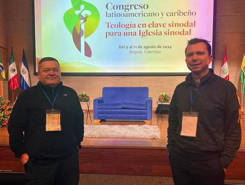 Dos hombres se encuentran de pie frente a un escenario con un fondo que anuncia un congreso latinoamericano y caribeño sobre teología.