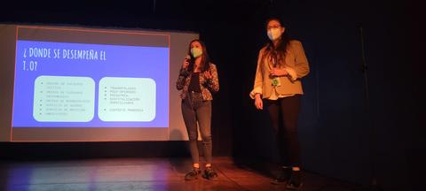 Dos presentadoras están hablando frente a una audiencia mientras se muestra una diapositiva en la pantalla.