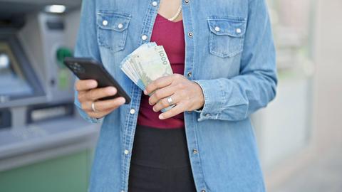 Una persona sostiene un teléfono móvil y billetes frente a un cajero automático.