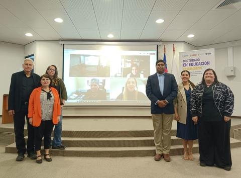 Un grupo de personas posando frente a una pantalla que muestra a otros participantes en videoconferencia durante un evento relacionado con la educación.