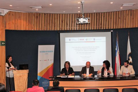 Una conferencia académica con un panel de expertos en una mesa junto a una presentación en pantalla.