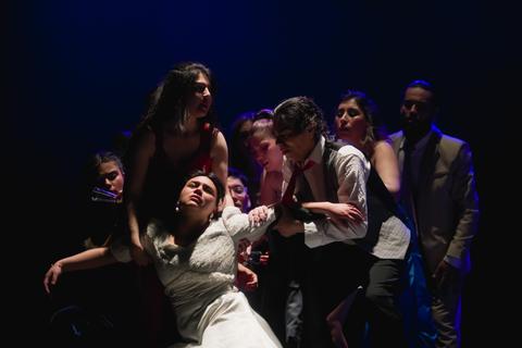 Un grupo de personas en un escenario, en una escena dramática donde una mujer vestida de blanco parece estar en peligro mientras otros la rodean.