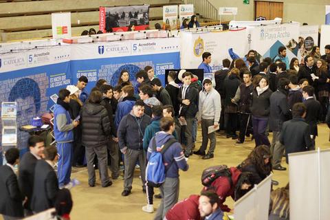 Un grupo de jóvenes se reúne en una feria educativa donde se exhiben diferentes stands de universidades y organizaciones.