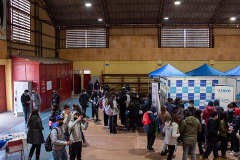 La imagen muestra un evento de estudiantes en un gimnasio, con puestos de información y numerosos asistentes interactuando.