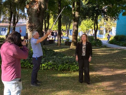 En la imagen se observa a una mujer posando mientras dos hombres interactúan, uno con una cámara y otro sosteniendo un objeto alargado.