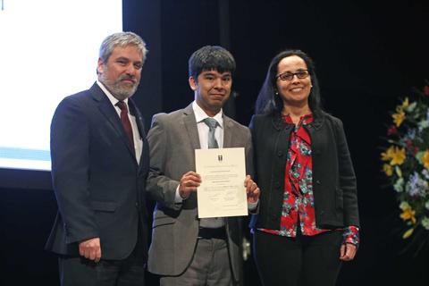 Un joven recibe un certificado en un evento formal acompañado de dos adultos.