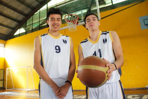 Dos jóvenes jugadores de baloncesto posan en una cancha con una canasta al fondo.