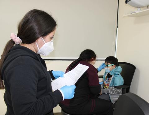 Una profesional de la salud está atendiendo a un niño en una consulta médica mientras otra persona revisa un documento.