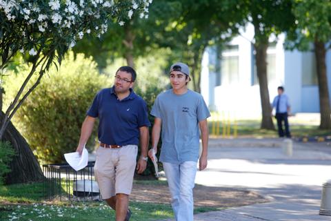 Dos hombres caminan juntos por un camino al aire libre, rodeados de árboles y flores.