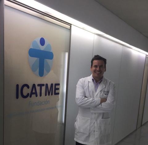 Un hombre sonriente viste un bata de doctor frente a una oficina con el logo de una fundación.