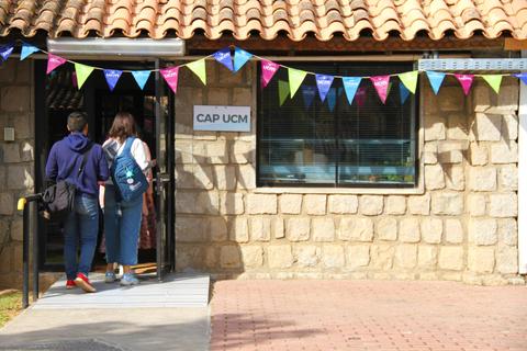 Un par de personas entra a un edificio con un letrero que dice 'CAP UCM' y un decorado de banderines de colores.
