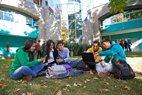 Un grupo de estudiantes sentados en el césped, utilizando dispositivos electrónicos y colaborando entre ellos frente a un edificio moderno.