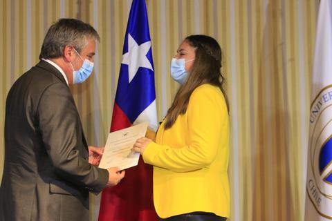 Un hombre y una mujer se entregan un certificado en un acto oficial frente a banderas.