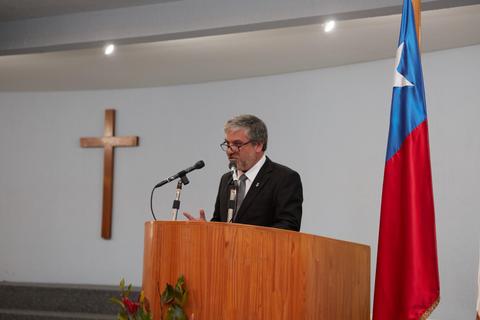 Un hombre de traje habla en un podio frente a una cruz y una bandera chilena.