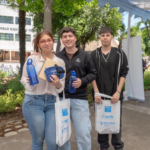 Tres jóvenes sostienen botellas de agua y otros objetos en un evento al aire libre.