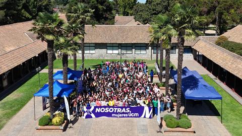 Un gran grupo de mujeres se reúne en un espacio abierto, frente a edificios con palmeras.