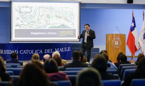 Un presentador habla sobre Magallanes y Elcano frente a una audiencia en un auditorio de la Universidad Católica del Maule.