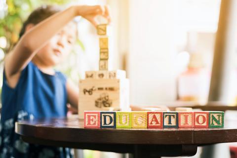 Una niña construye una torre con bloques de madera en una mesa, mientras que la palabra 'EDUCATION' está formada con otros bloques en la superficie.