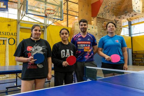 Cuatro jóvenes posan en una sala de deportes con mesas de ping pong y un ambiente de competencia amistosa.