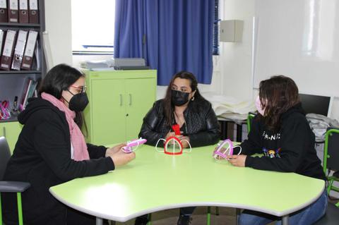 Tres mujeres sentadas alrededor de una mesa verde, usando mascarillas y sosteniendo objetos en las manos.