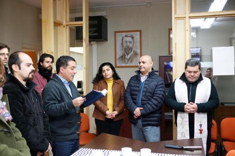 Un grupo de personas asiste a una ceremonia mientras un hombre lee un libro en un entorno formal.