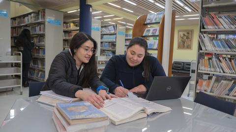 Dos estudiantes están trabajando juntas en una biblioteca, revisando libros y tomando apuntes.