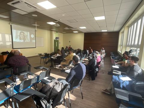 Una sala de clases con varias personas sentadas en bancos mirando hacia una pantalla.