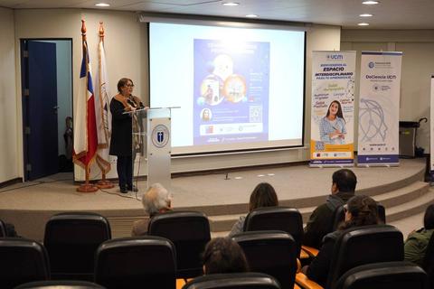 Una mujer habla en un podio durante un evento en un auditorio con banderas y pantallas informativas al fondo.