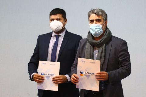 Dos hombres con mascarillas sosteniendo documentos frente a un fondo neutro.