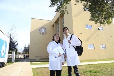 Dos estudiantes con batas de laboratorio posan frente a un edificio educativo.