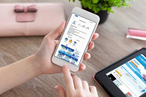 Una persona sostiene un teléfono móvil mostrando una aplicación, mientras que en la mesa hay una tablet y una planta decorativa.