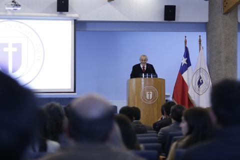 Un hombre habla desde un podio en una conferencia, con banderas chilenas y una pantalla detrás.