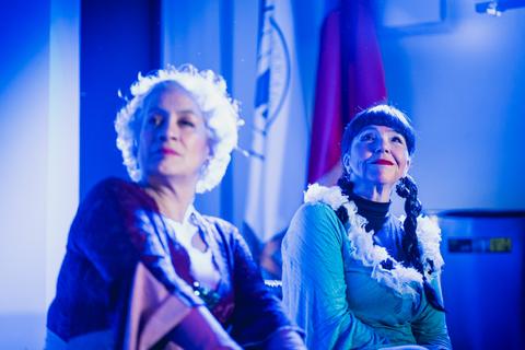 Dos mujeres sentadas, con vestimenta colorida y sonriendo en un ambiente iluminado por luces azules.