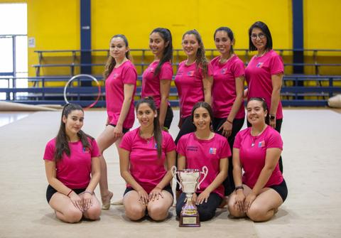 Un grupo de gimnastas en un gimnasio muestran un trofeo mientras posan en equipo.