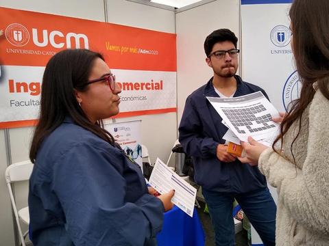 Dos personas conversan en un stand de la Universidad Católica del Maule sobre la carrera de Ingeniería Comercial.