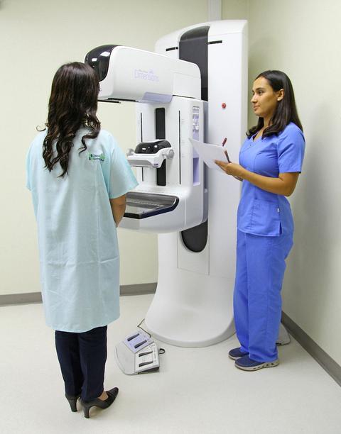Una mujer en bata se prepara para una mamografía mientras una profesional de la salud le explica el procedimiento.
