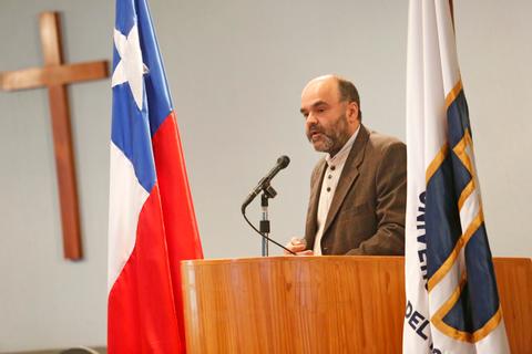 Un hombre habla desde un podio mientras se encuentran presentes la bandera de Chile y un símbolo de una universidad.