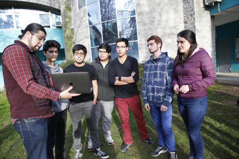 Un grupo de jóvenes observa a un hombre que les muestra algo en una computadora portátil en un exterior con césped.