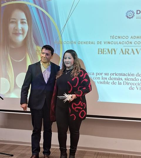 Una pareja posa para la foto con un fondo que muestra un reconocimiento o premio.