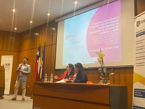 Un hombre presenta una charla en un auditorio mientras dos mujeres lo escuchan y se encuentran frente a él.