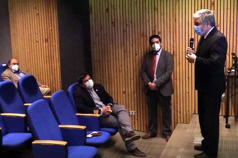 Un hombre habla en un micrófono mientras otros lo escuchan en una sala con sillas azules.