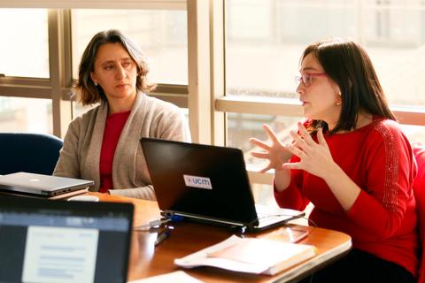 Dos mujeres están participando en una reunión, una de ellas habla gesticulando mientras la otra escucha atentamente.