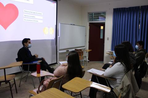 Un grupo de estudiantes escucha a un docente en un aula mientras se proyecta una presentación en la pantalla.