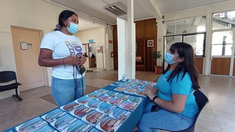 Dos mujeres conversan en un espacio con una mesa llena de folletos informativos.