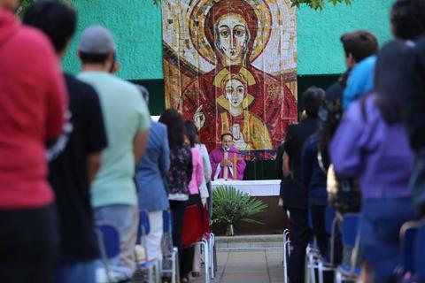 Un grupo de personas asiste a una ceremonia religiosa frente a un altar con un fondo de arte religioso.