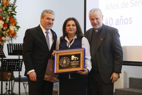 La imagen muestra a tres personas en un evento, una mujer sostiene un reconocimiento entre dos hombres.